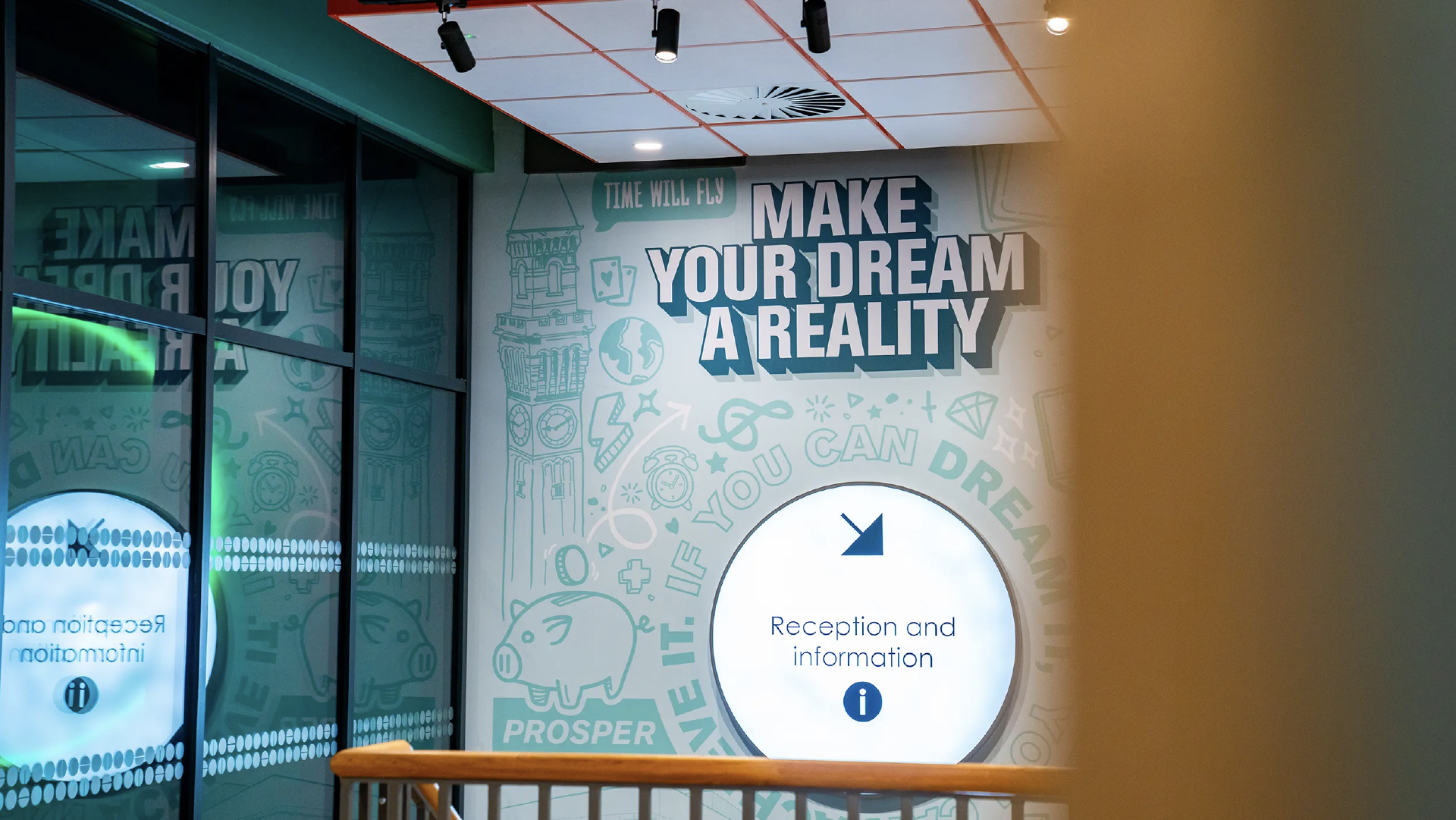Reception wall at Pritchatt's House Hub reading 'Make your dream a reality' in bold type, over teal line illustrations of Old Joe, a piggy bank and alarm clocks, beside a circular reception and information sign