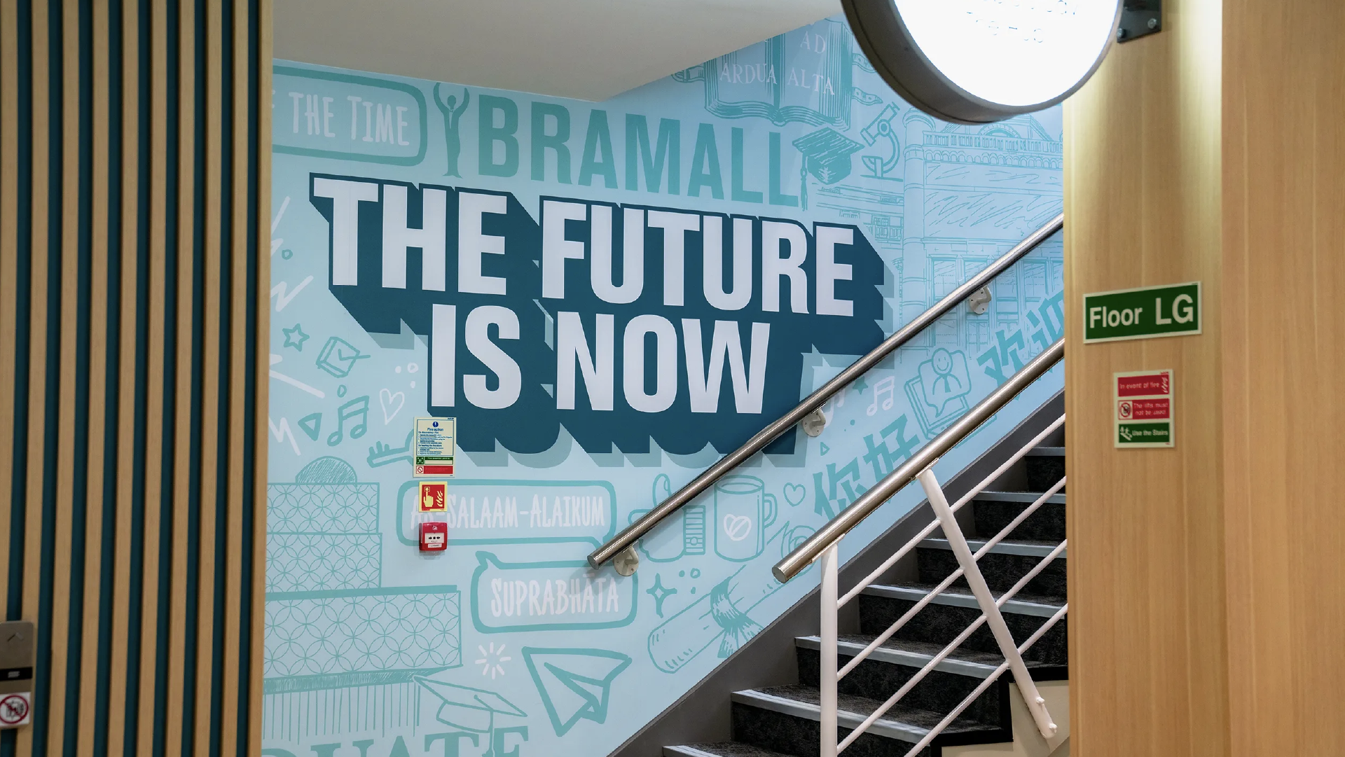 Installed stairwell mural reading 'The future is now' in white and navy over a pale blue illustrated wall, beside the staircase
