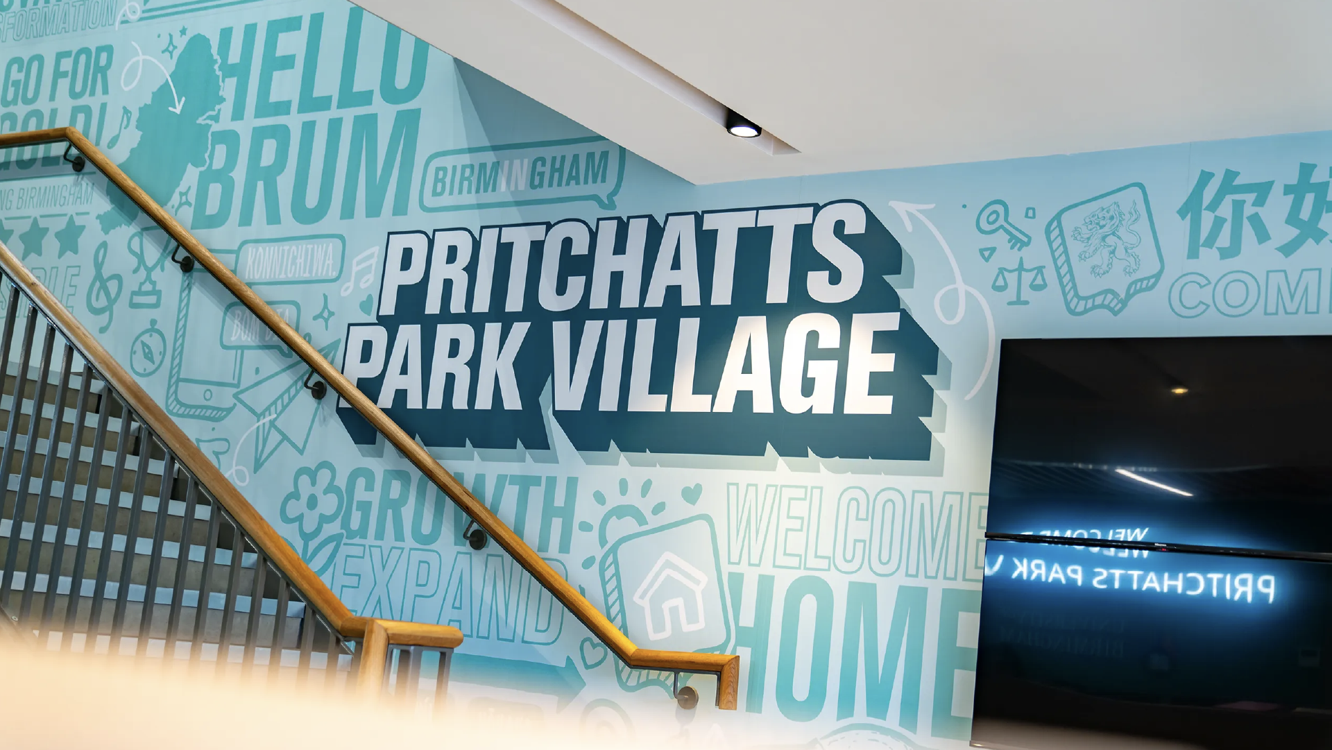 The 'Pritchatts Park Village' stairwell graphic with a timber handrail, next to a digital screen displaying a welcome message